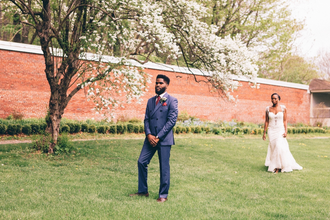 A groom looking towards the camera just as the bride walks up to him from behind for the first look moment in a garden  