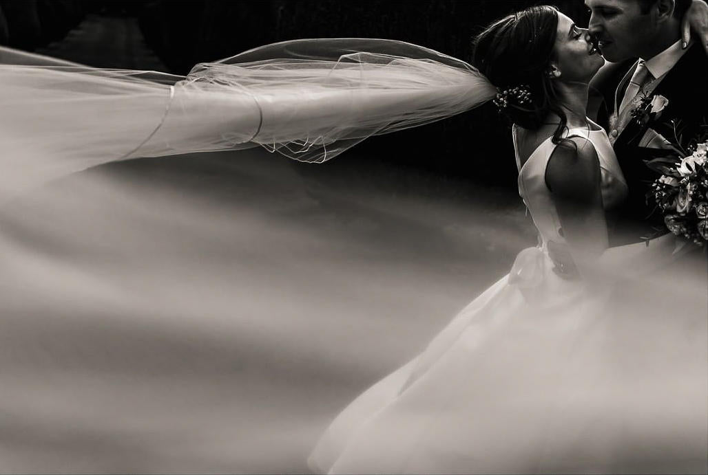 A bride's veil blowing in the wind after the ceremony — kissing her husband. Photo edited by ShootDotEdit.