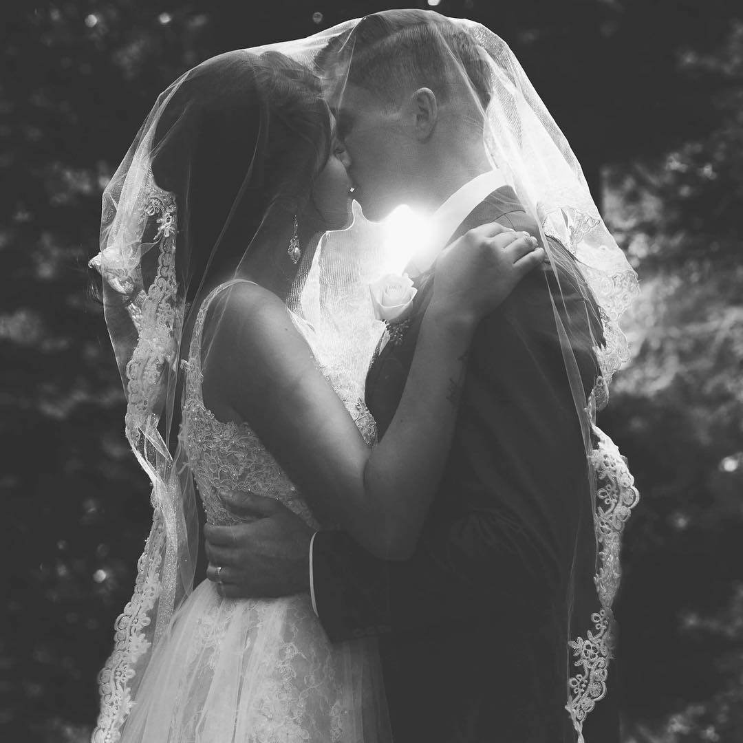 Black and white portrait of a bride and groom kissing while being covered by the veil