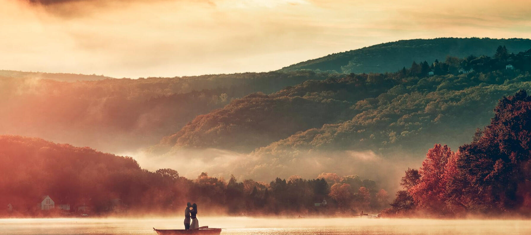 A couple posing on a boat in front of a hillside landscape colorfully lit by sun rays