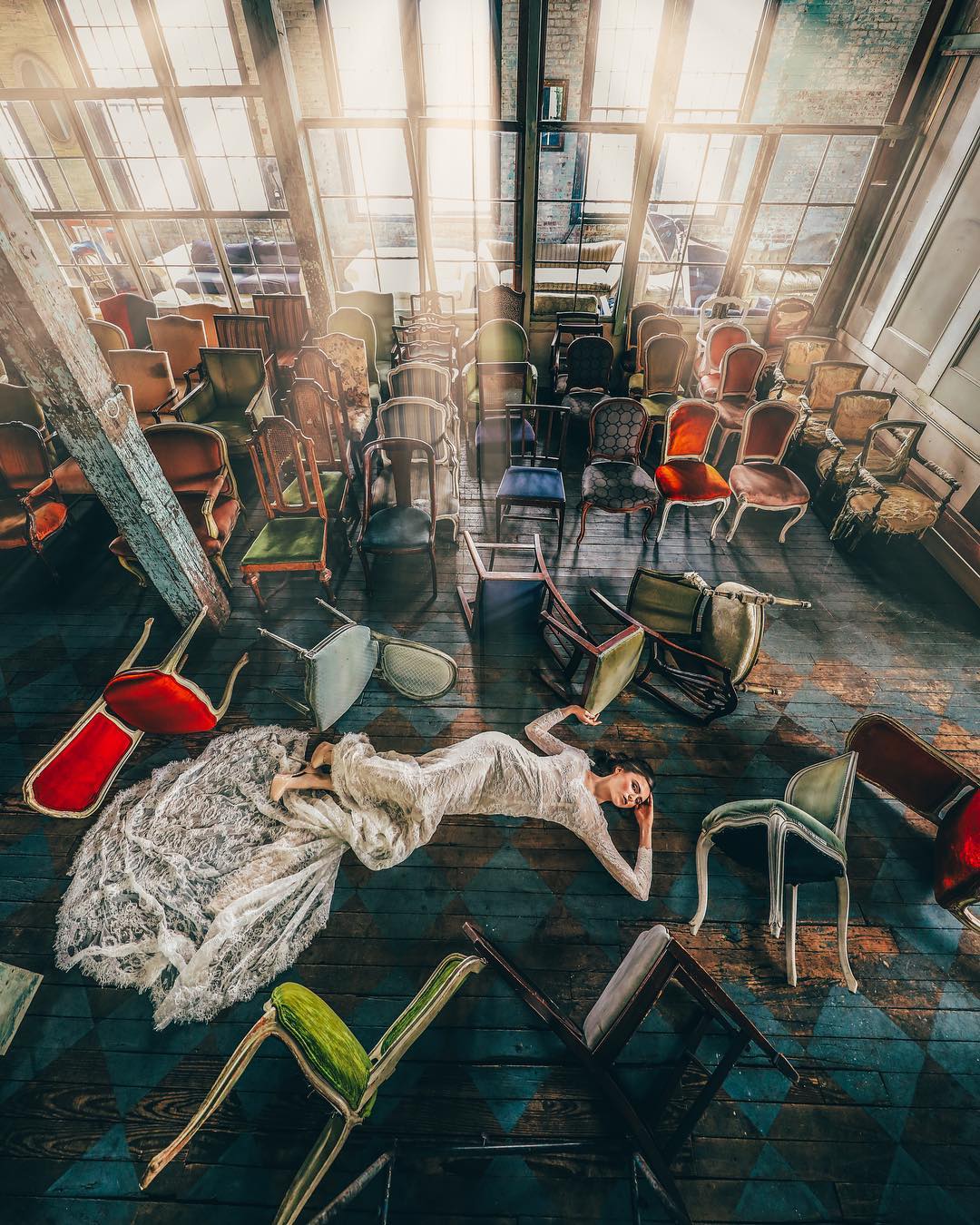 Artistic shot of a bride lying on the floor surrounded by fallen chairs in a room that has sunlight gushing from the windows