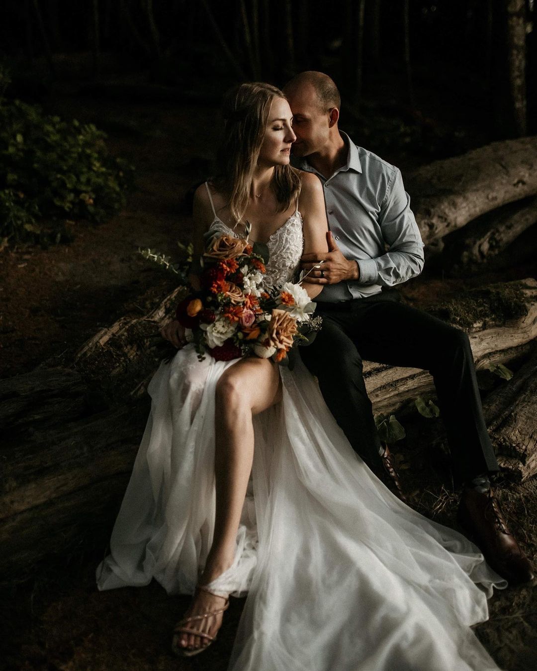 A bride and groom posing facing each other while being seated on a log 