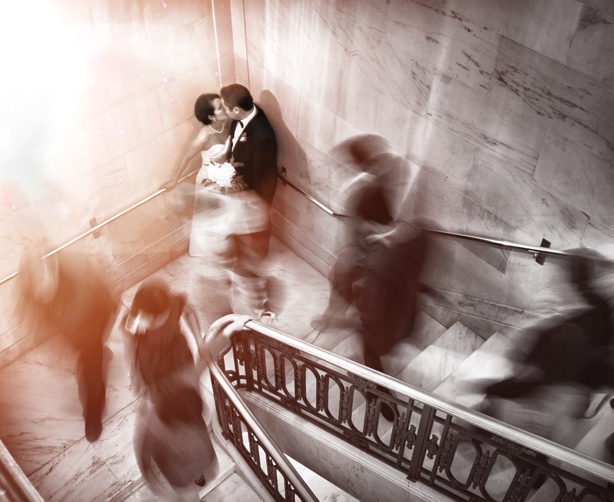 A bride and groom kissing at the corner of a stairway as the guests are captured in a blur as they walk around 