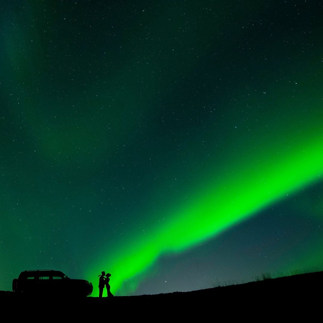 Silhouette of a couple looking at each other alongside a car under the sky full of stars and northern lights