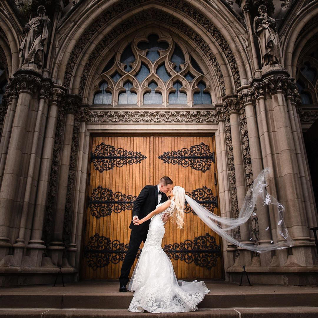 A bride an groom embracing on cathedral steps while the bride's veil flows in the wind.