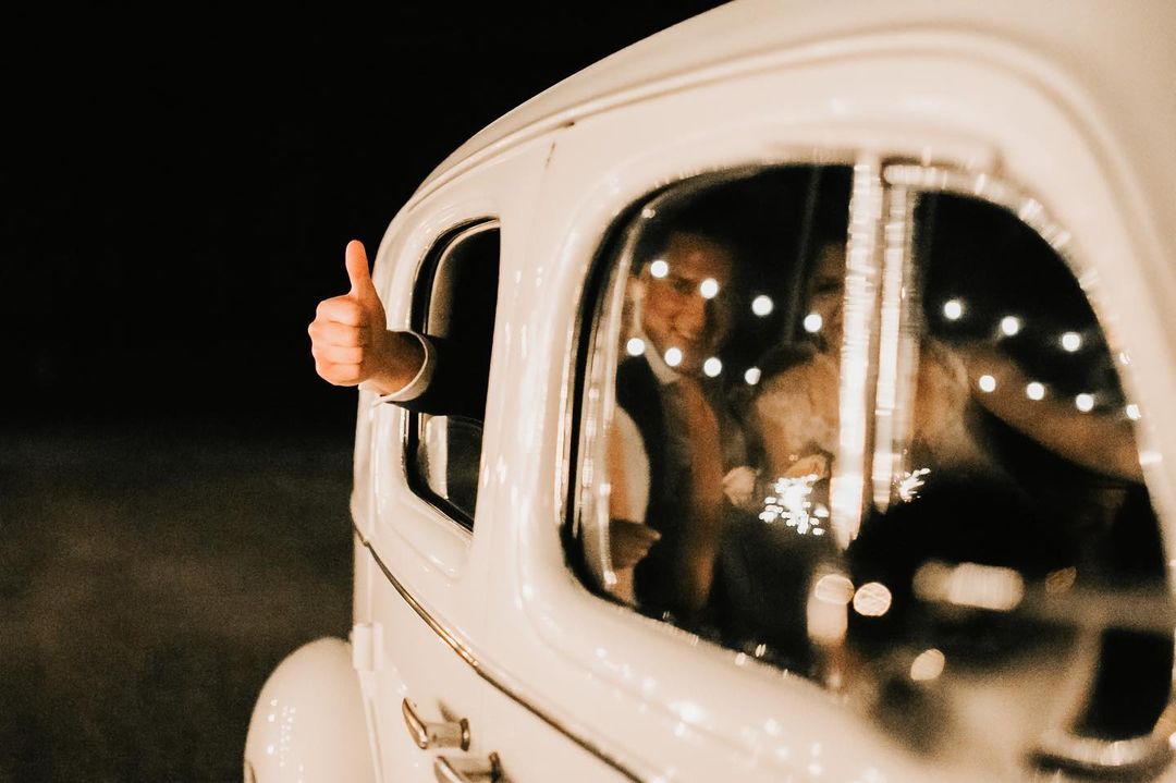 Bride and groom seated inside a white car for making the exit with groom showing a thumbs up from the car's window 