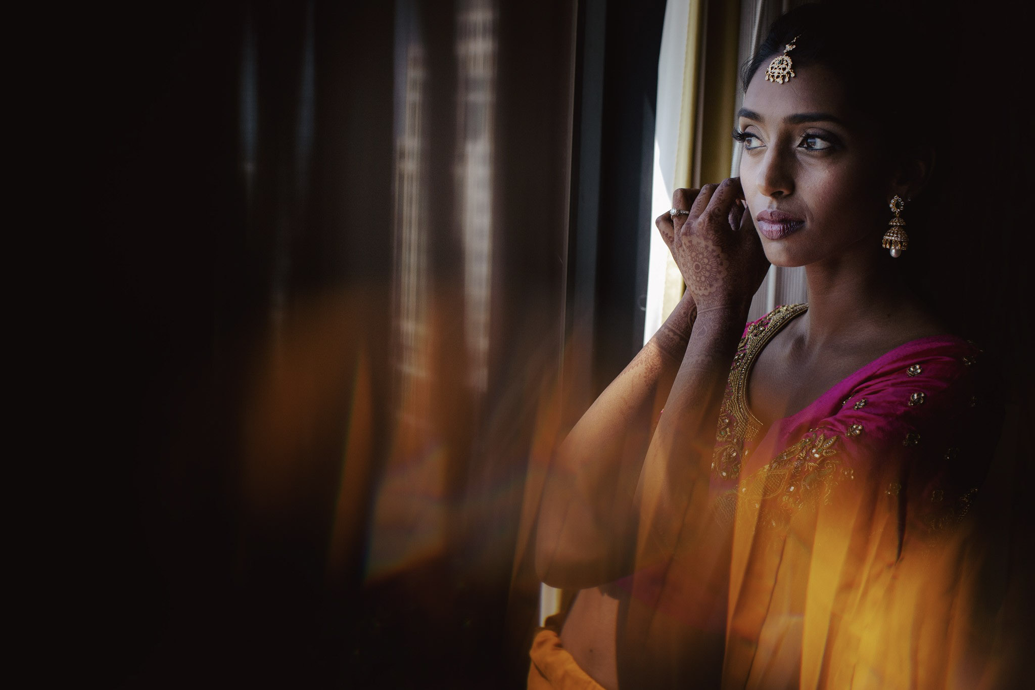 Mid angle shot of an Indian bride getting ready and wearing earrings in front of a window