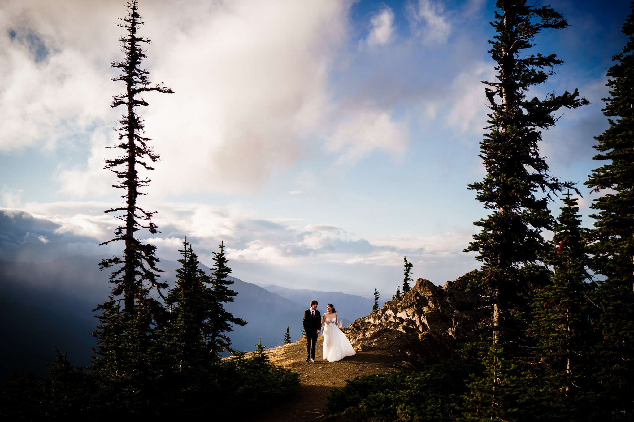 A bride and groom holds hands on top of a mountain top with cloud-covered mountain peaks at the background