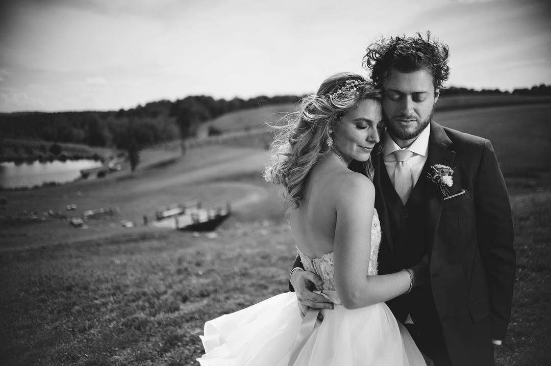 black and white image of a woman wearing flower crown with eyes closed leaning against man wearing suite on their wedding day in an open field near a lake