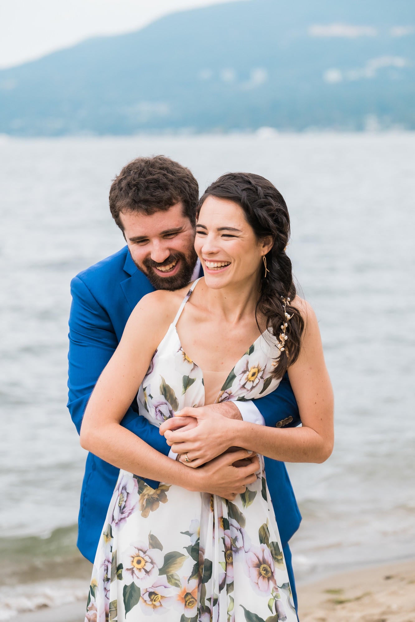 couple leaning into each other and laughing on the edge of the water with a hill behind them