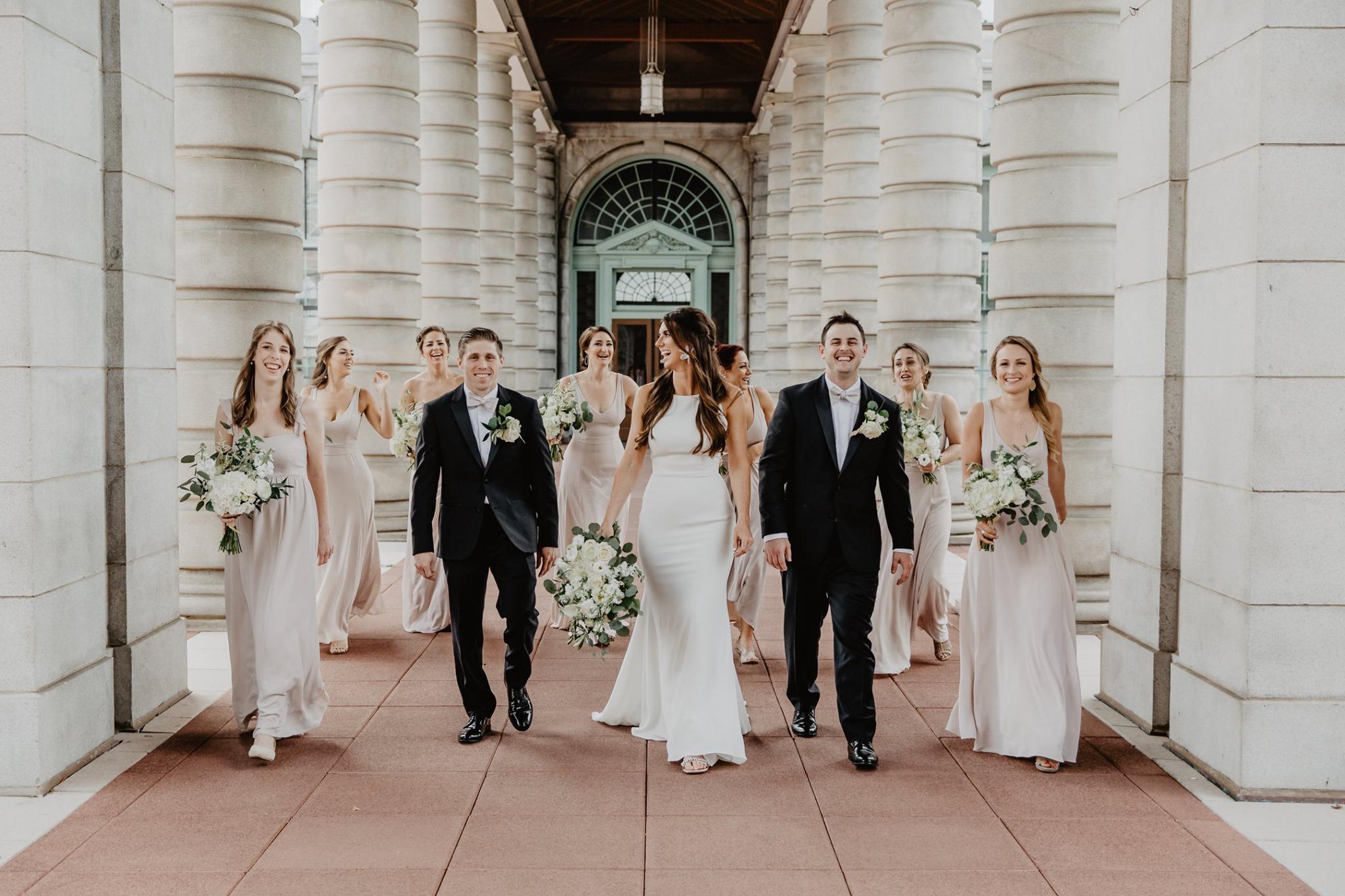 bride, groom and wedding party walking towards photographer, laughing, with white columns on either side