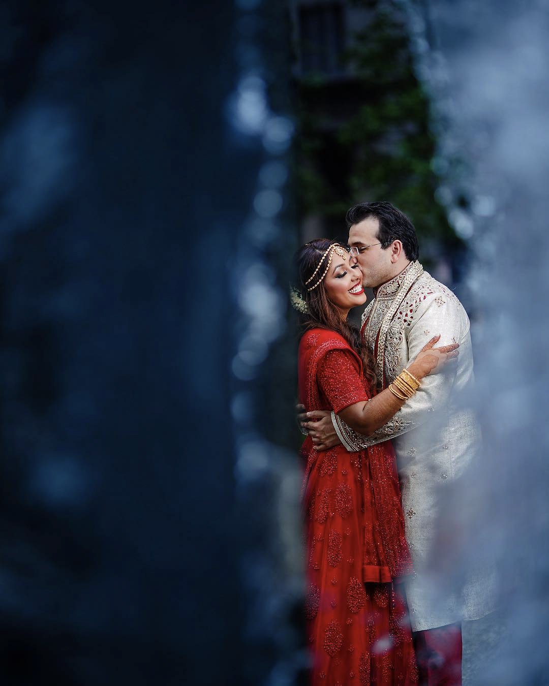 Bride and groom dressed in traditional Indian wedding clothing posing for the camera as they hug and kiss each other