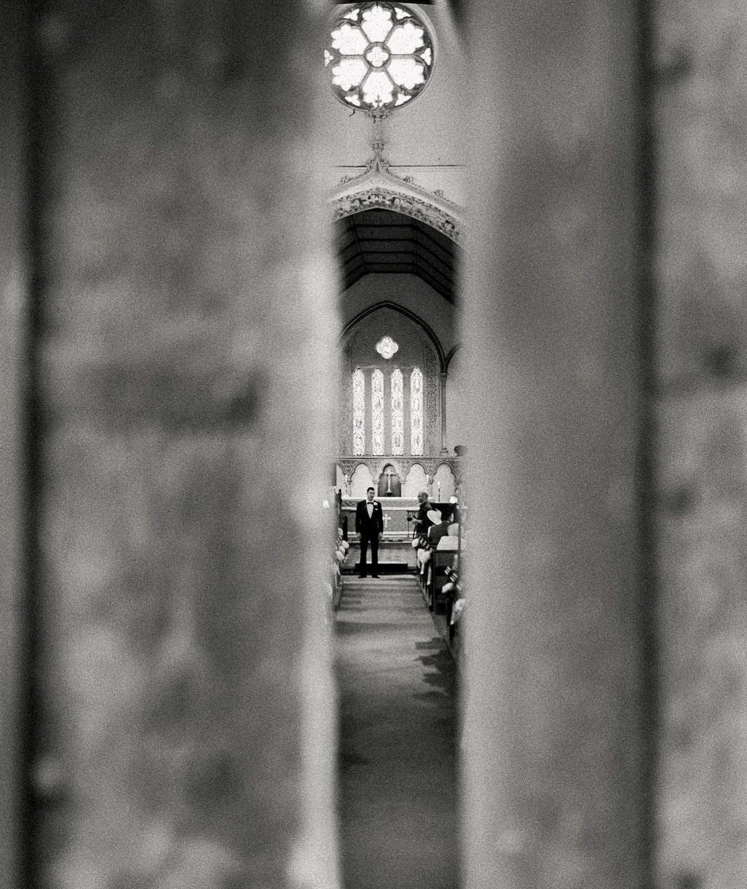 a black and white image through a gap of a groom waiting for the bride at the altar