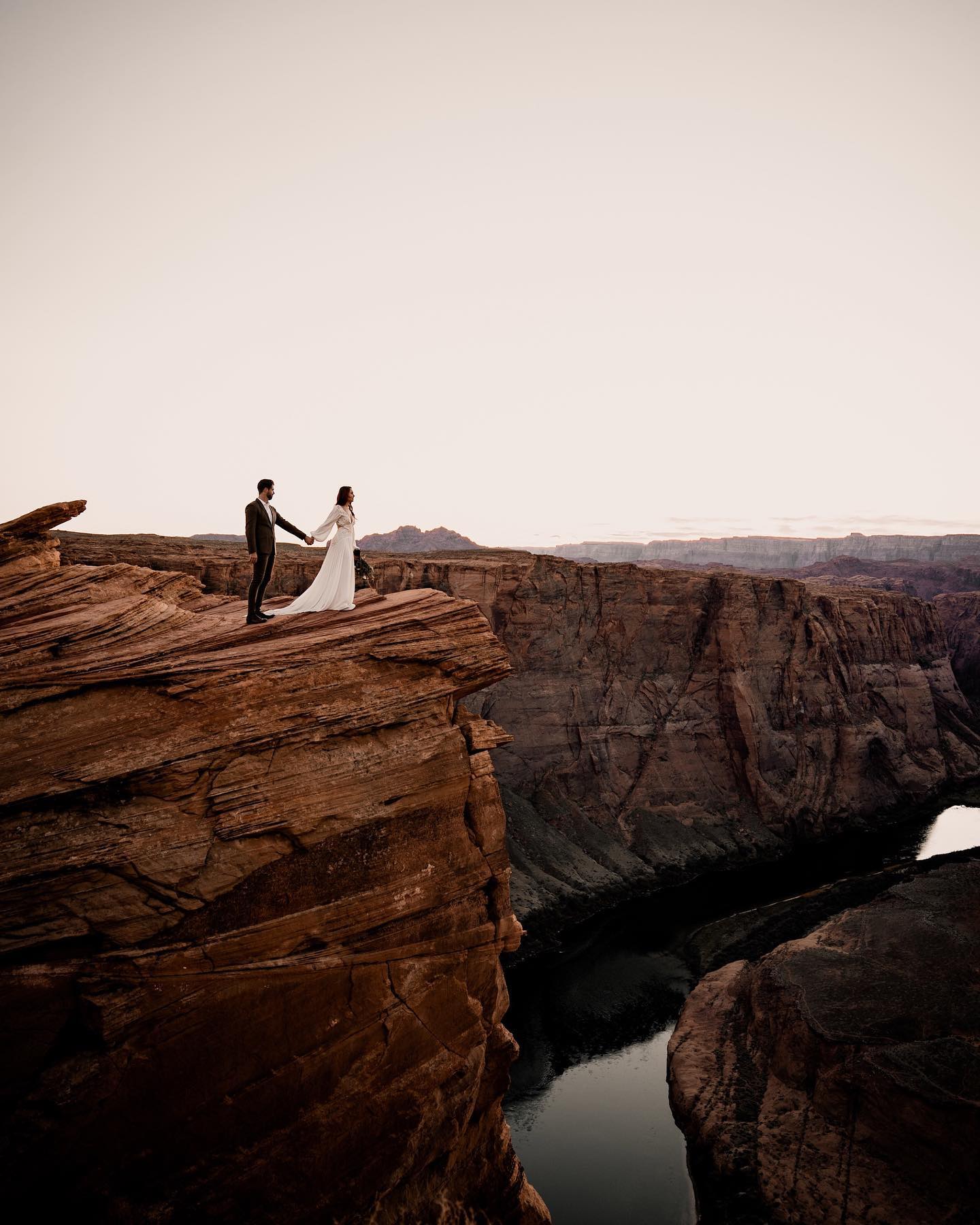 a wedding couple holding hands and standing at the edge of a cliff overlooking a river