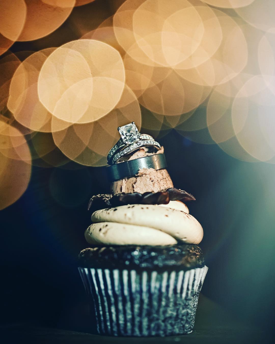 Two wedding rings placed on top of a chocolate cupcake with cream frosting with a bokeh effect background