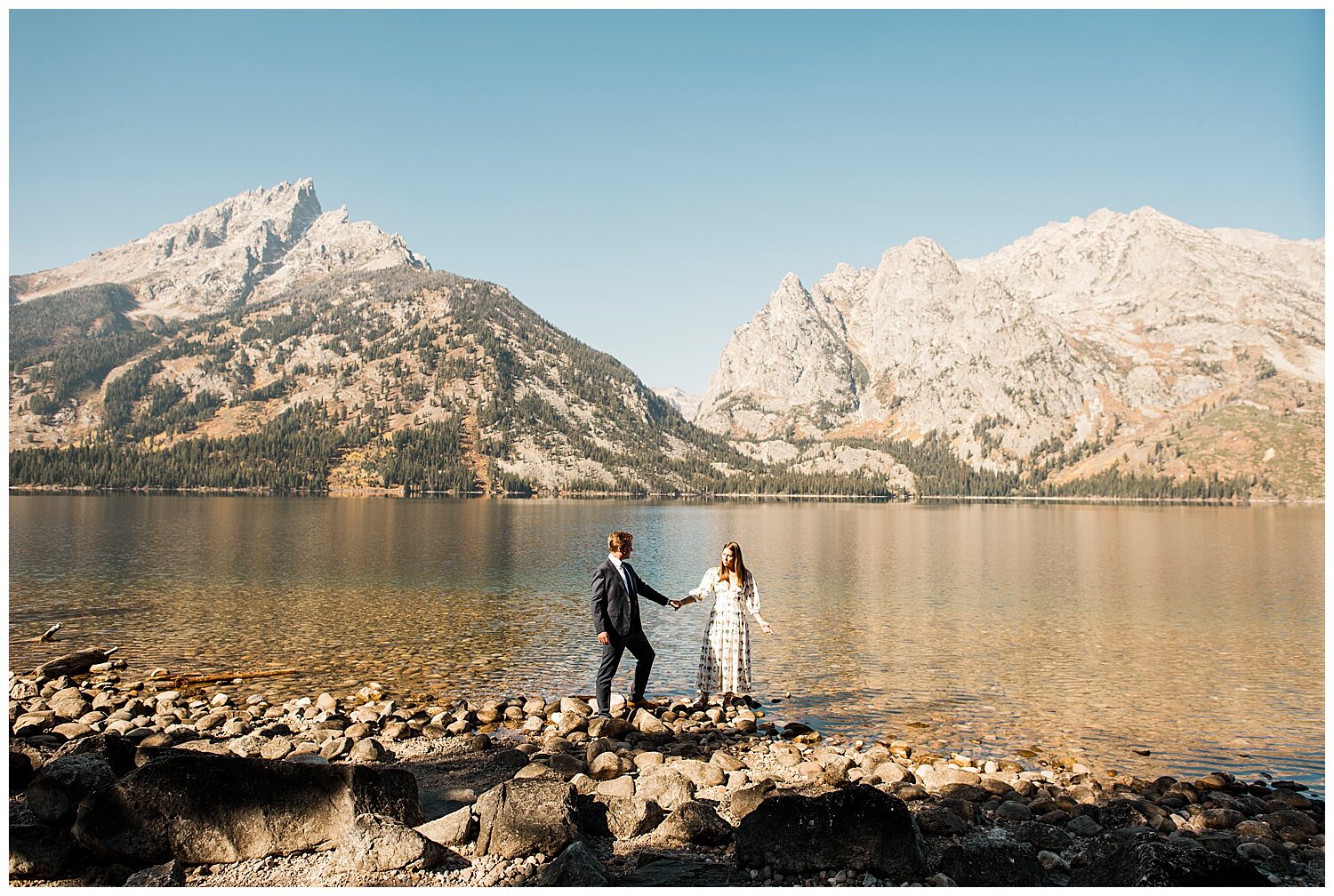 A couple holding hands while posing at the side of a lake with mountains at the background