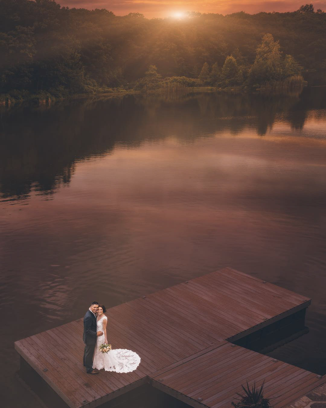 a wedding couple standing on a pier by a lake while the sun is setting