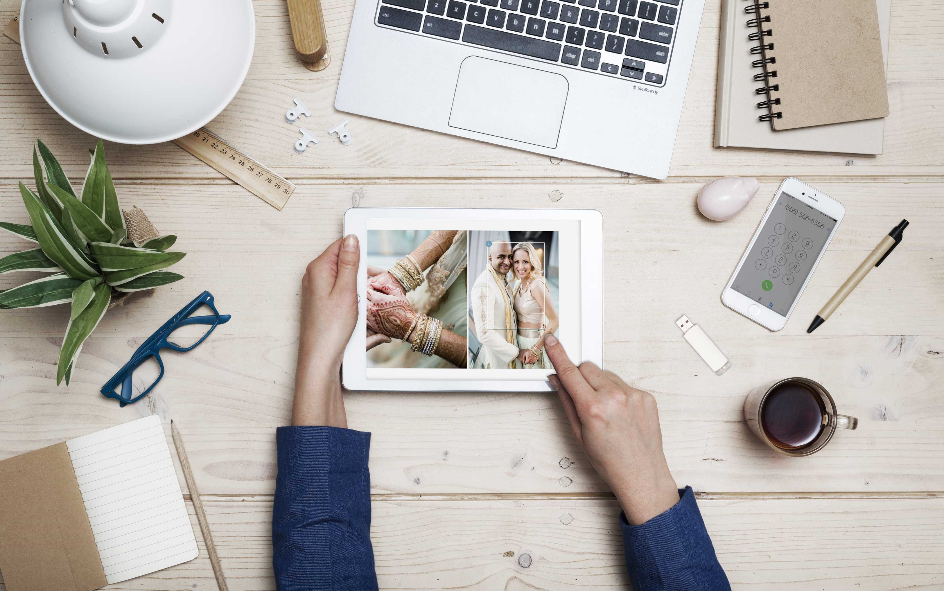 A close up of a wooden desk showcasing human hands showing pictures in a digital tablet that is surrounded by other things