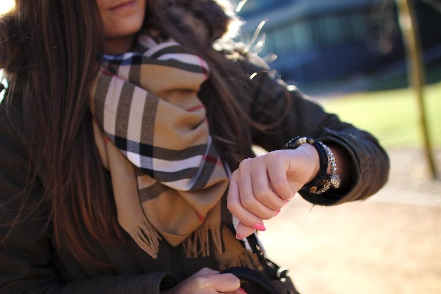 An image of a woman with a brown scarf and jacket as she looks down at her watch, which is on her arm along with a silver bracelet.