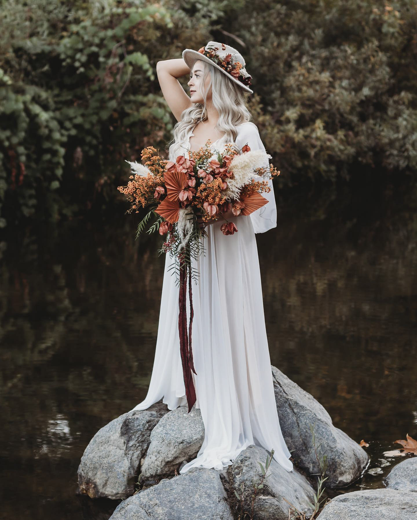 a bride to be standing on some rocks by a lake posing with the bridal bouquet