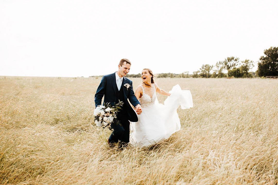 couple in wedding attire running in gold field and laughing while groom carries bouquet