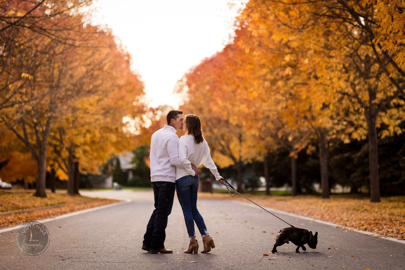 a couple kissing with their pet around in the middle of the street surrounded by trees 