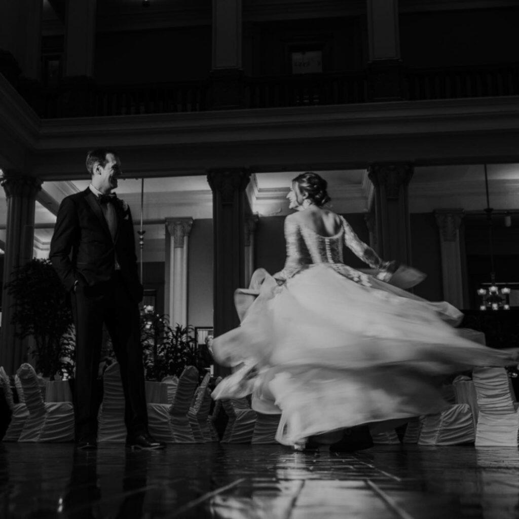 a black and white image of a bride twirling in her wedding dress while the groom smiles at her