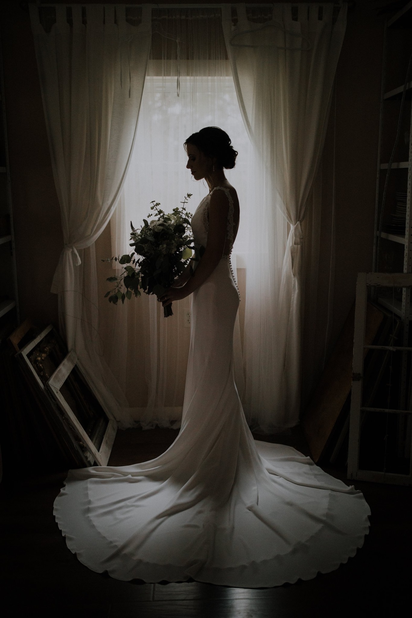 A silhouette of a bride posing in front of a window as she holds a bridal bouquet 