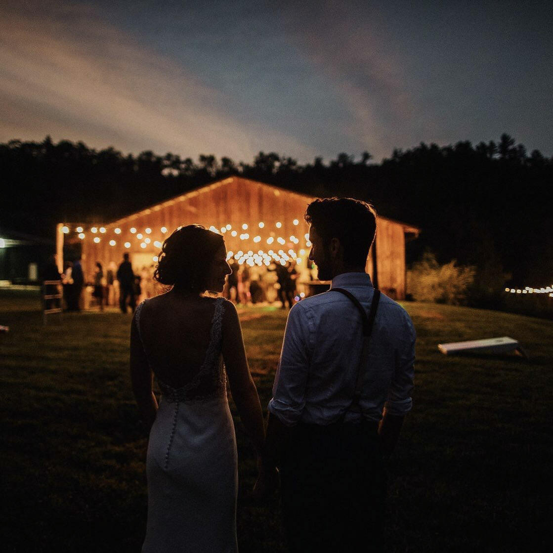 a couple standing holding hands in front of a blurred lit up wedding reception