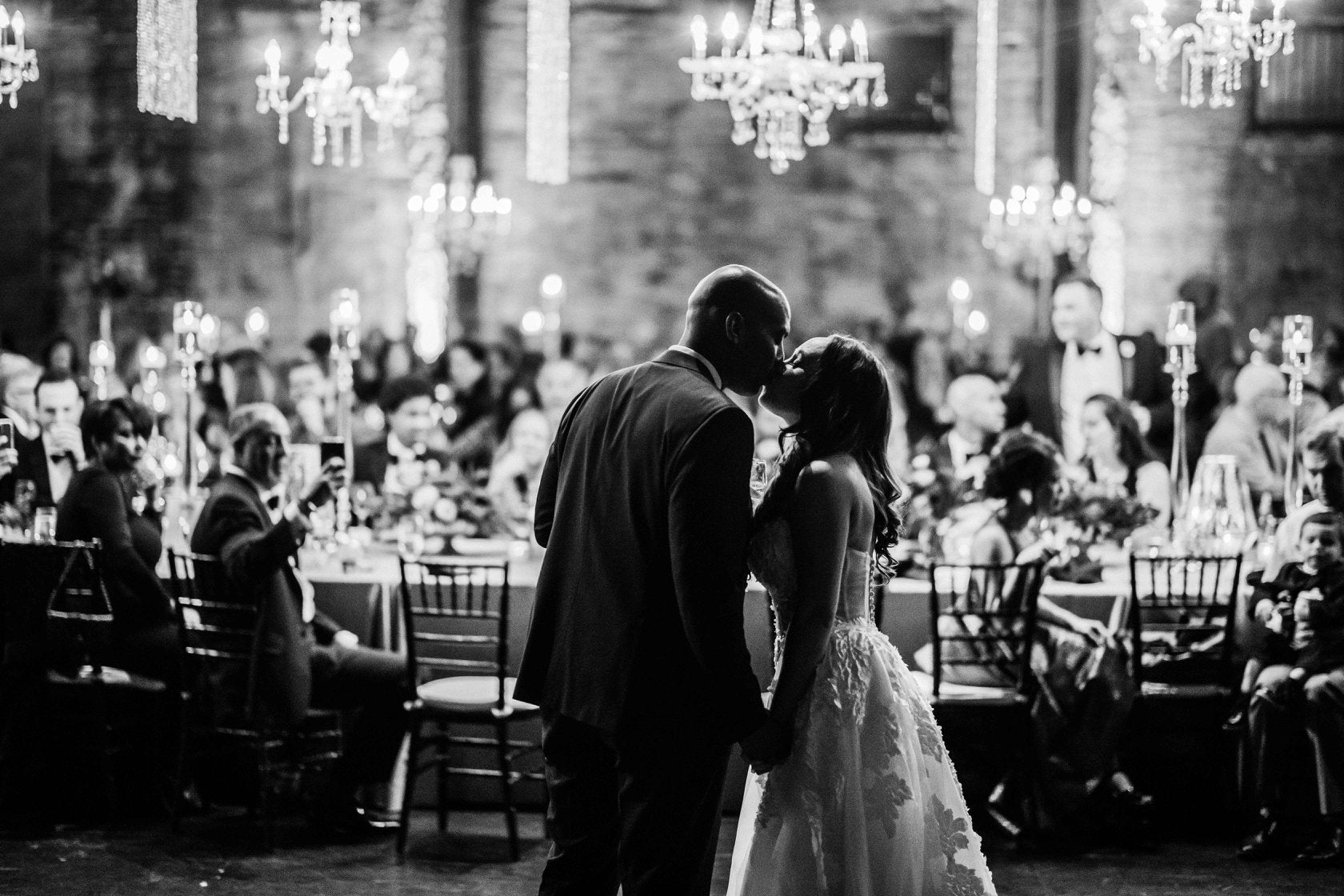 Black and white image of the bride and groom hold hand and kiss as the guests watch over from the dinner table