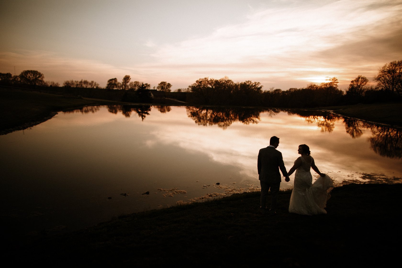 A silhouette of a bride and groom standing by the lakeside during sunset