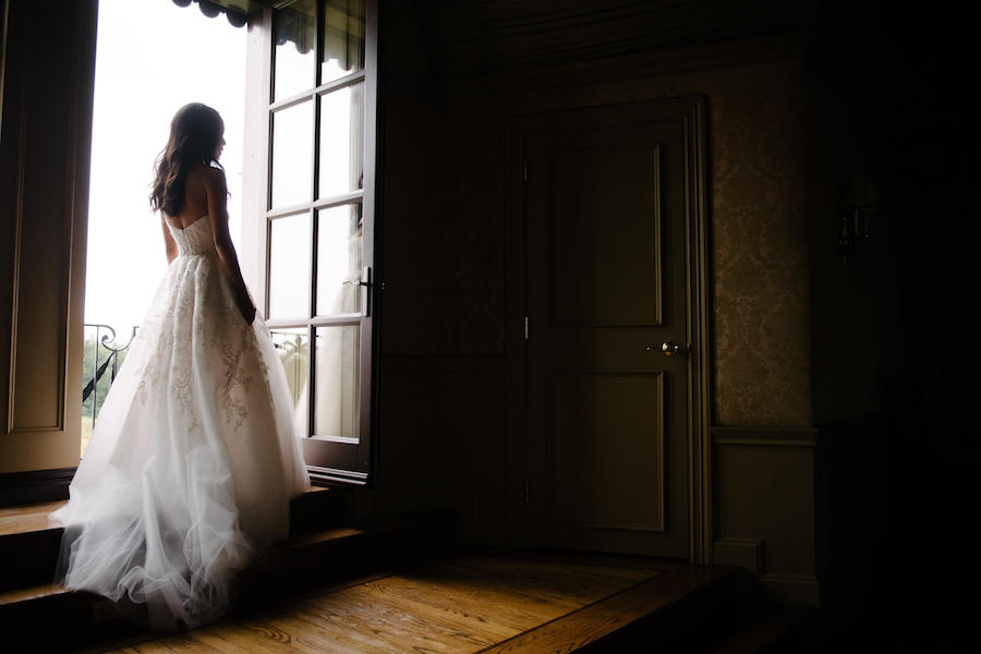 A wedding bridal portrait of the bride facing the door, her back to the camera and natural light coming in to highlight her and the details of her wedding dress.