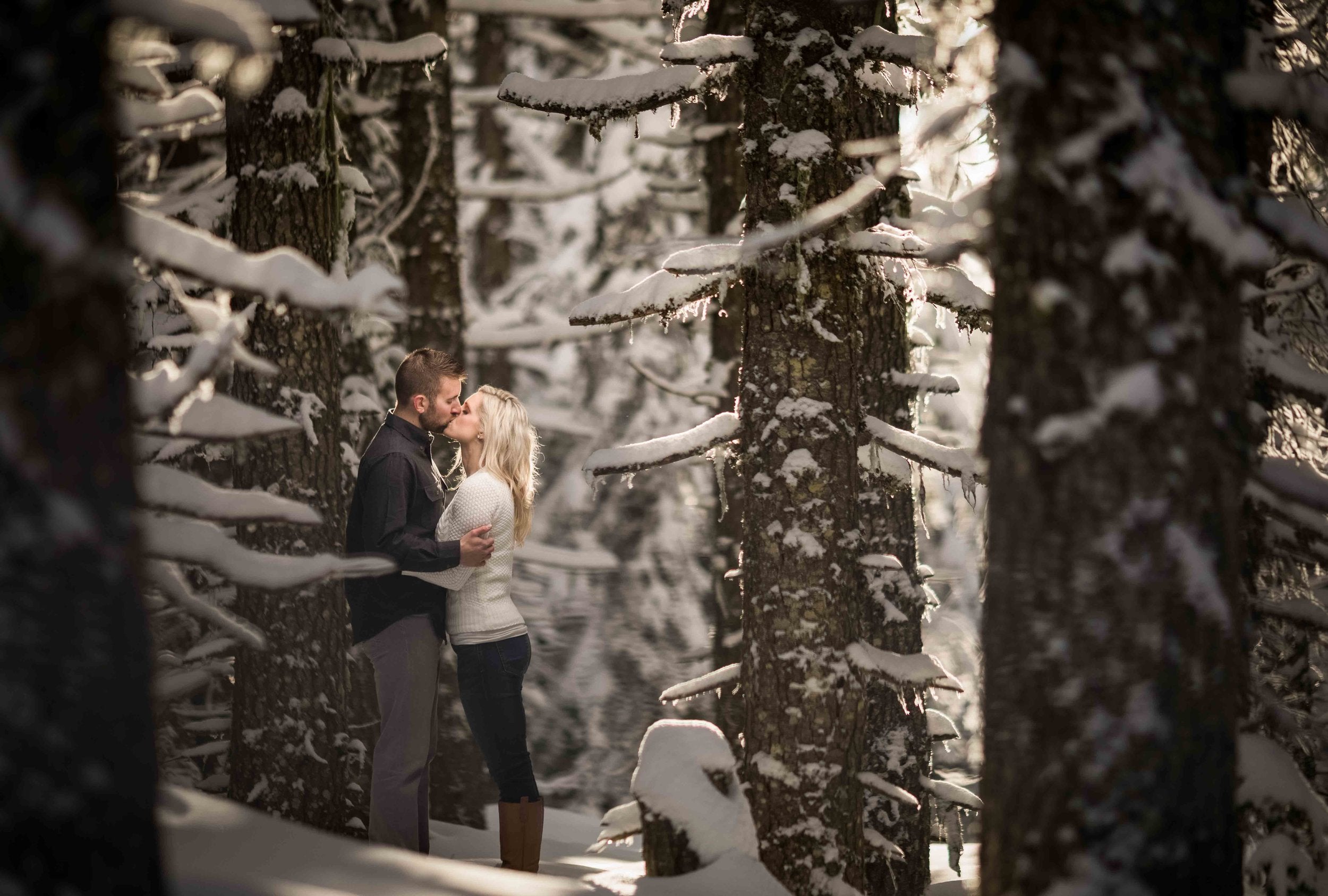 A couple kissing in the wilderness alongside snow-covered tree branches