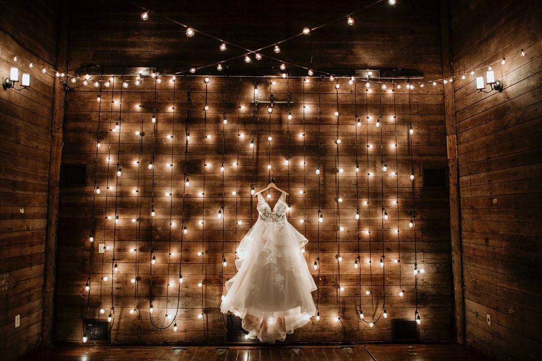 A wide-angle shot of a wedding dress hung on the wall with a brown background that is decorated with fairy lights