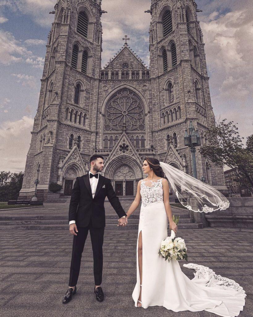 a newlywed bride and a groom standing holding each other's hands in front of a church