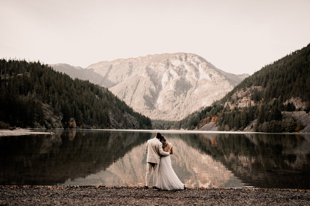 a newly wed couple standing in front a lake surrounded by mountains 