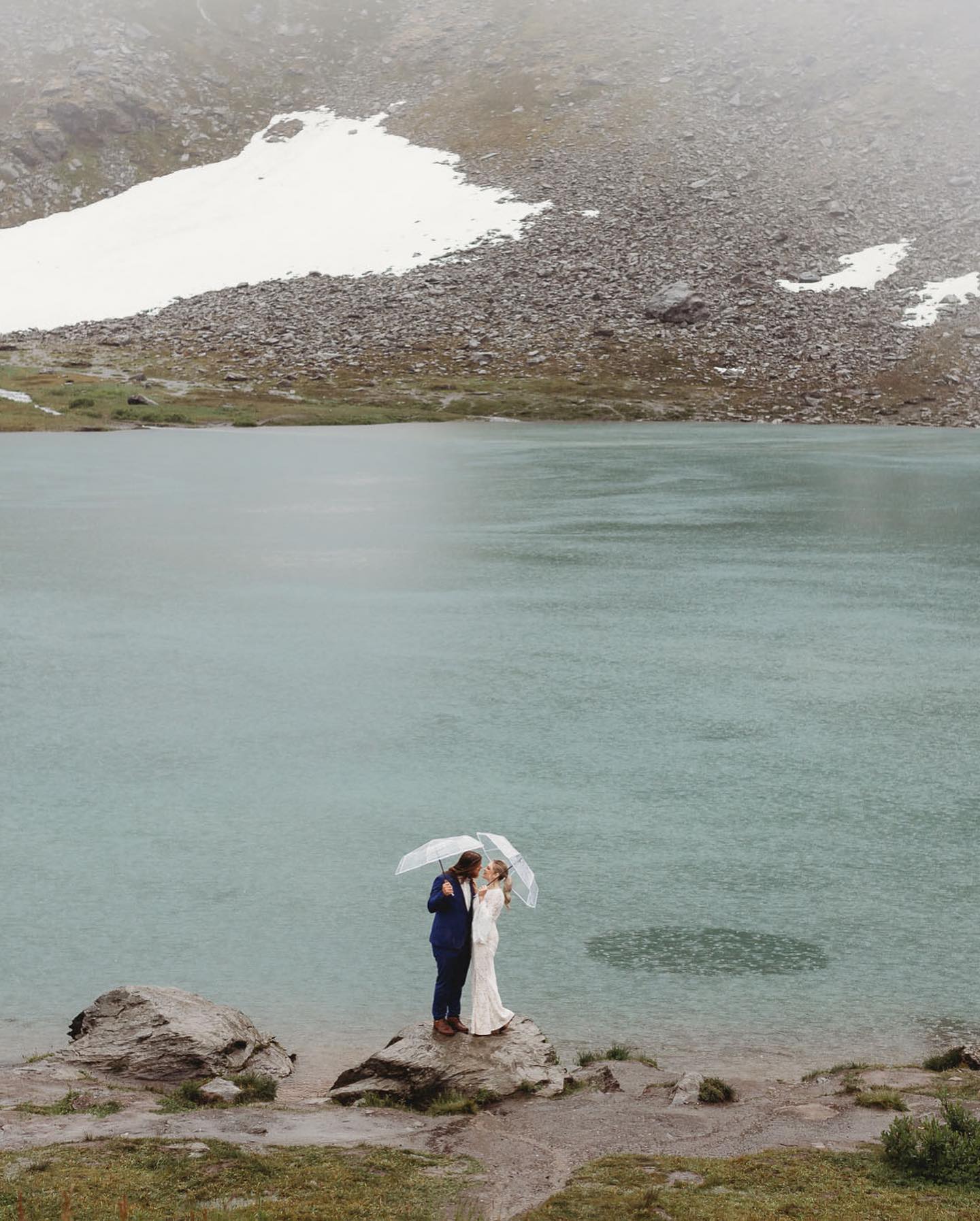 a couple standing by a lake in their wedding dress holding umbrellas while it is raining