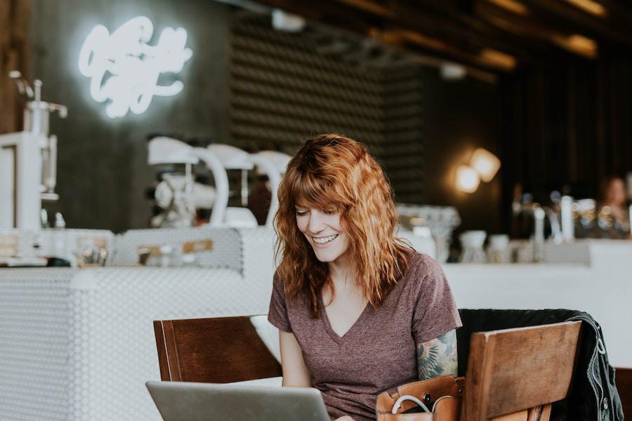 An image of a woman with red hair and arm tattoos in a coffee shop, smiling and looking down at the laptop in front of her.