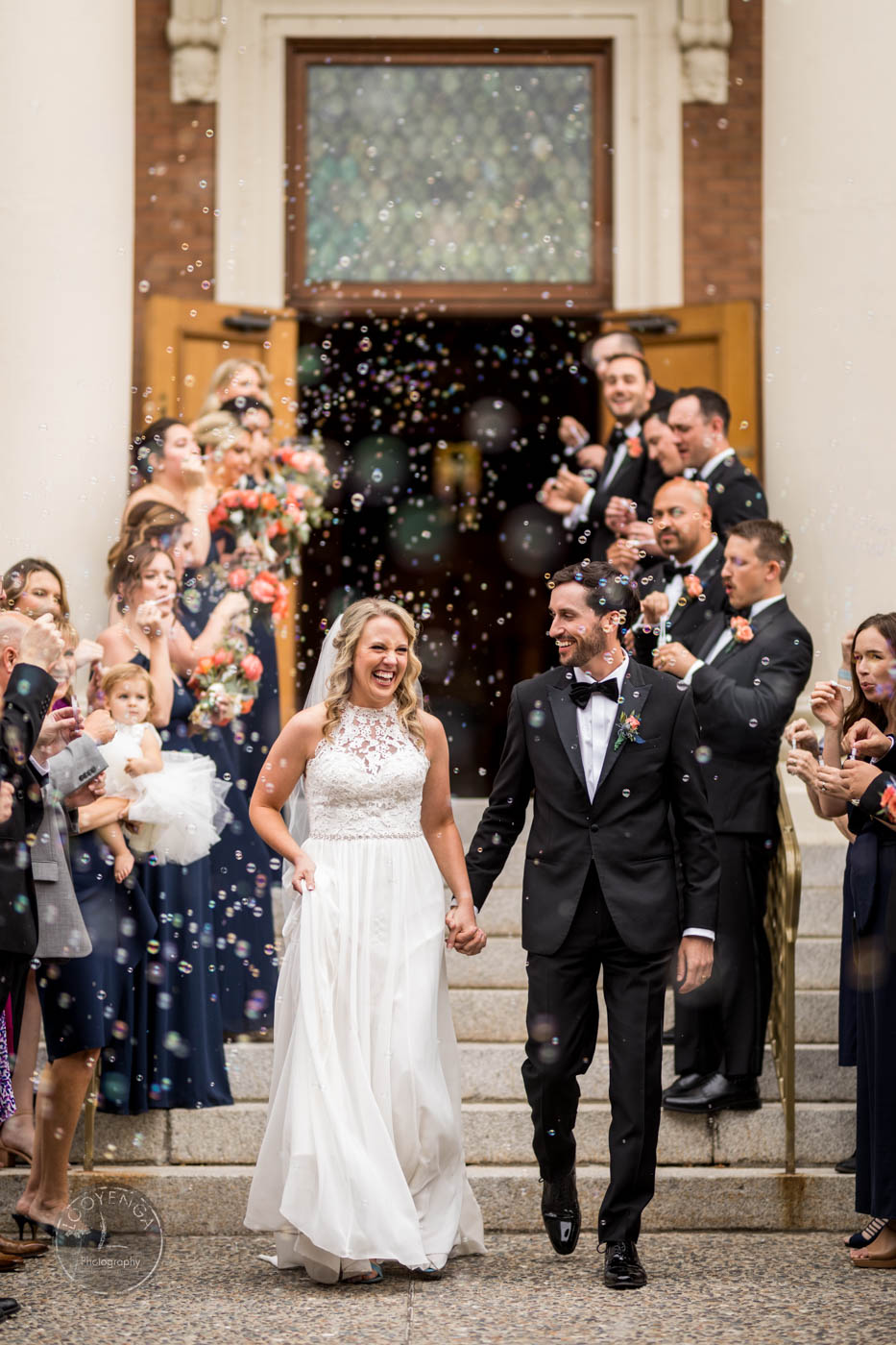 a newly wed couple exiting their wedding venue while the guests are blowing bubbles