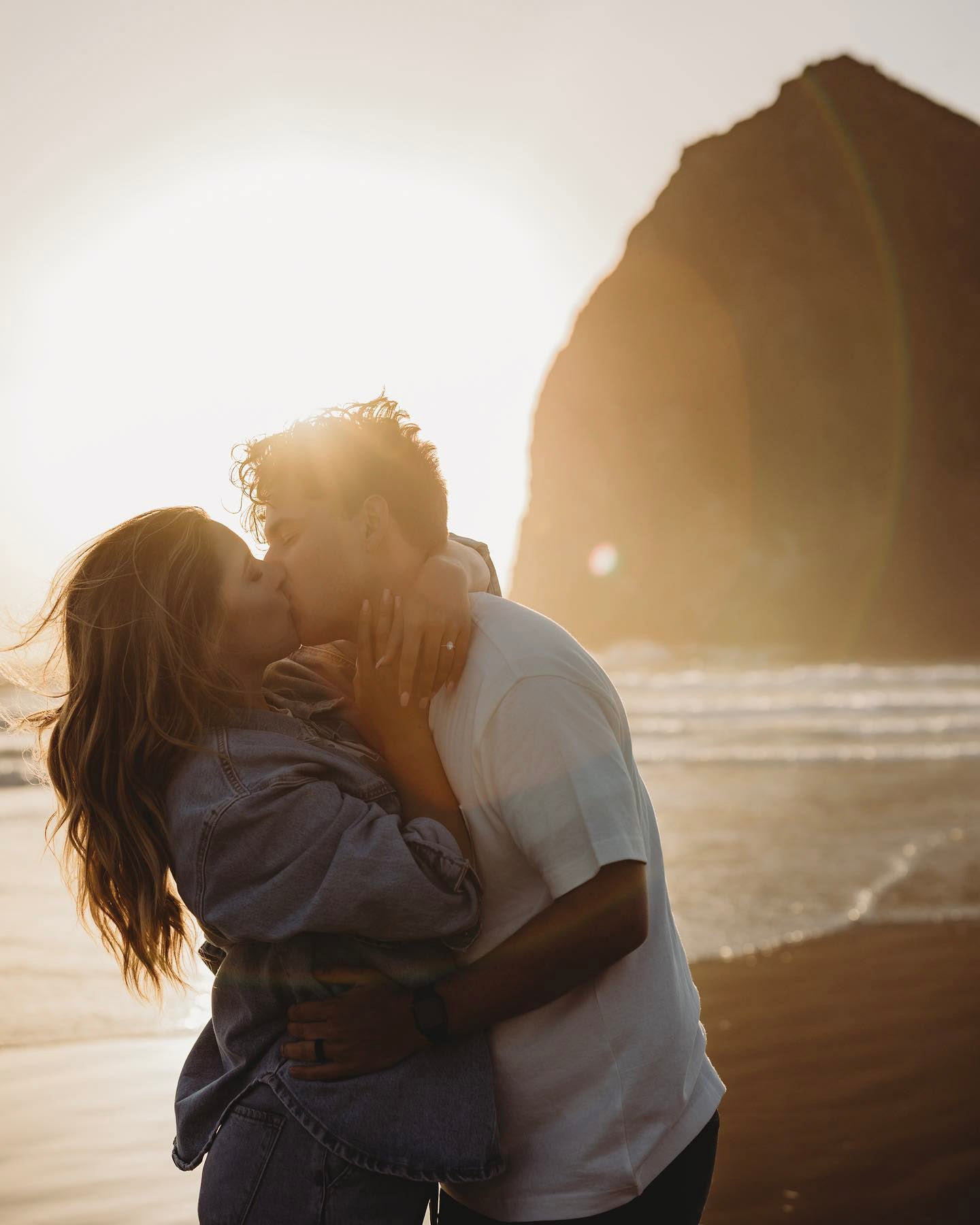 an engaged couple kissing on the beach while the sun shines behind them