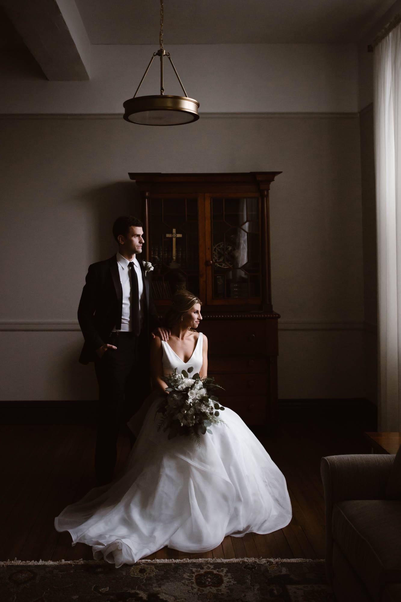 A bride and groom posing while sitting on a chair that is placed in front of a window