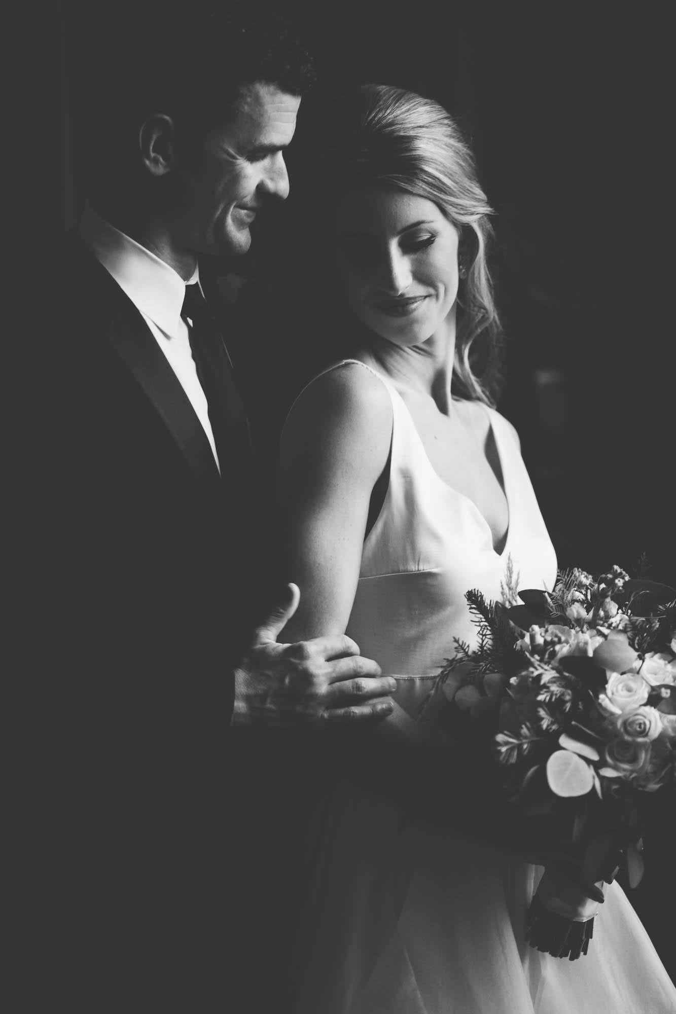 Black and white photo of a bride and groom posing in a dark room in front of a window