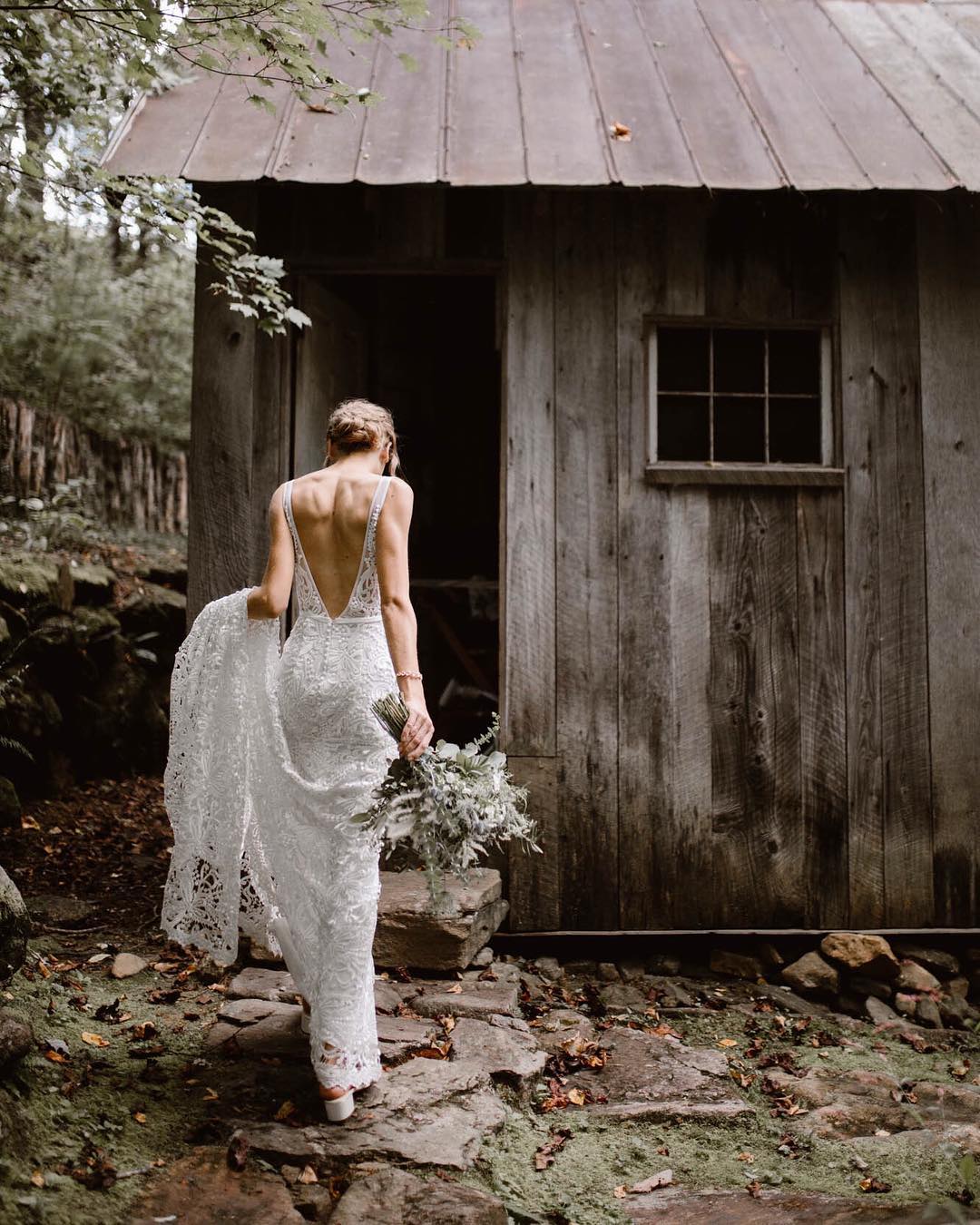 A bride holding the train of her dress from one hand and the bouquet from the other walks into a hut