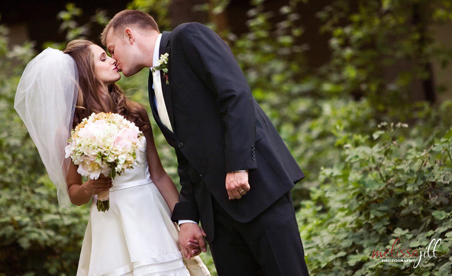 Bride and groom kissing, while the bride holds her bouquet in one hand and holds the groom's hand with the other.