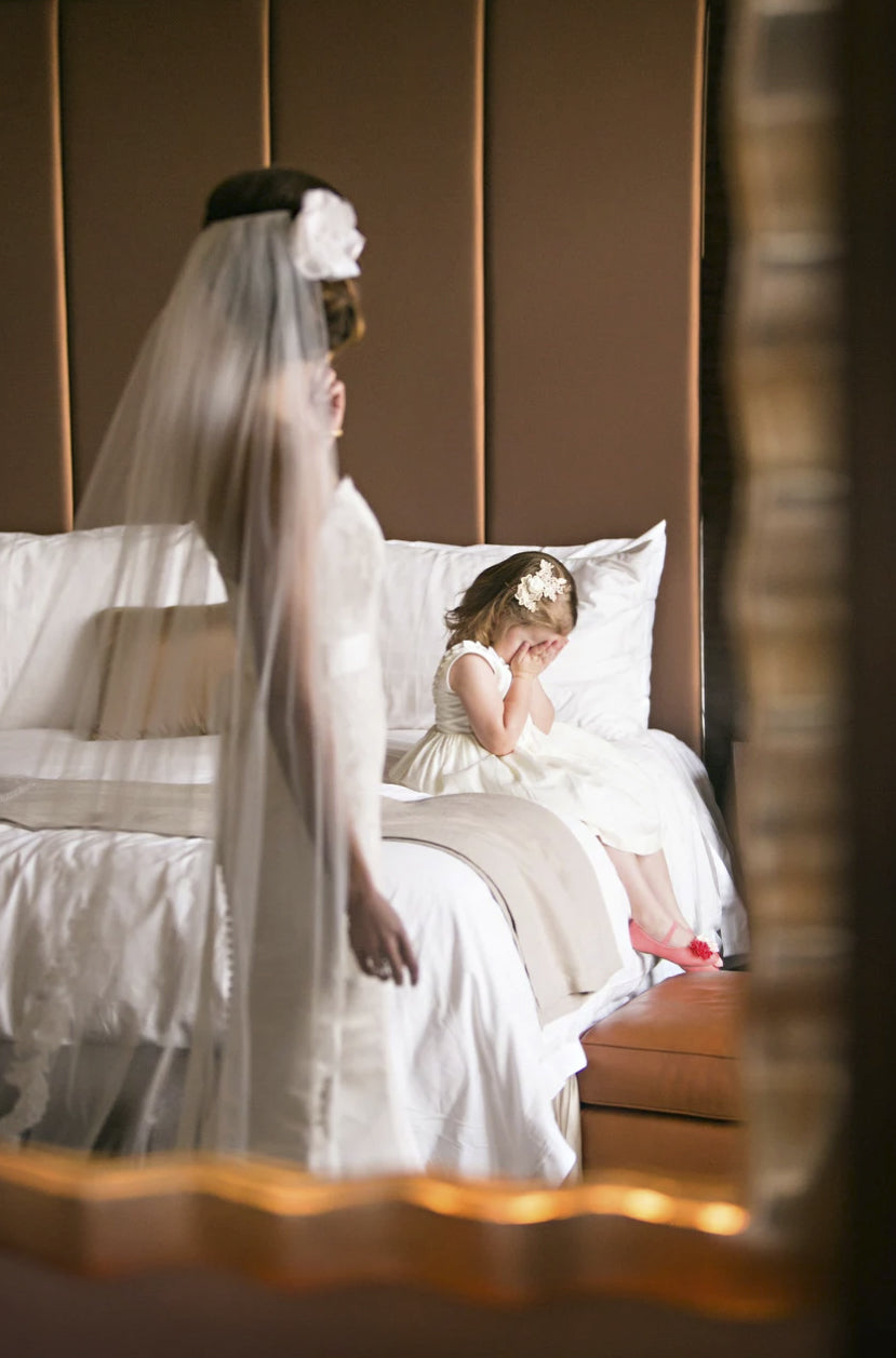 The bride watches over the flower girl sitting on the bed with hands placed over her eyes