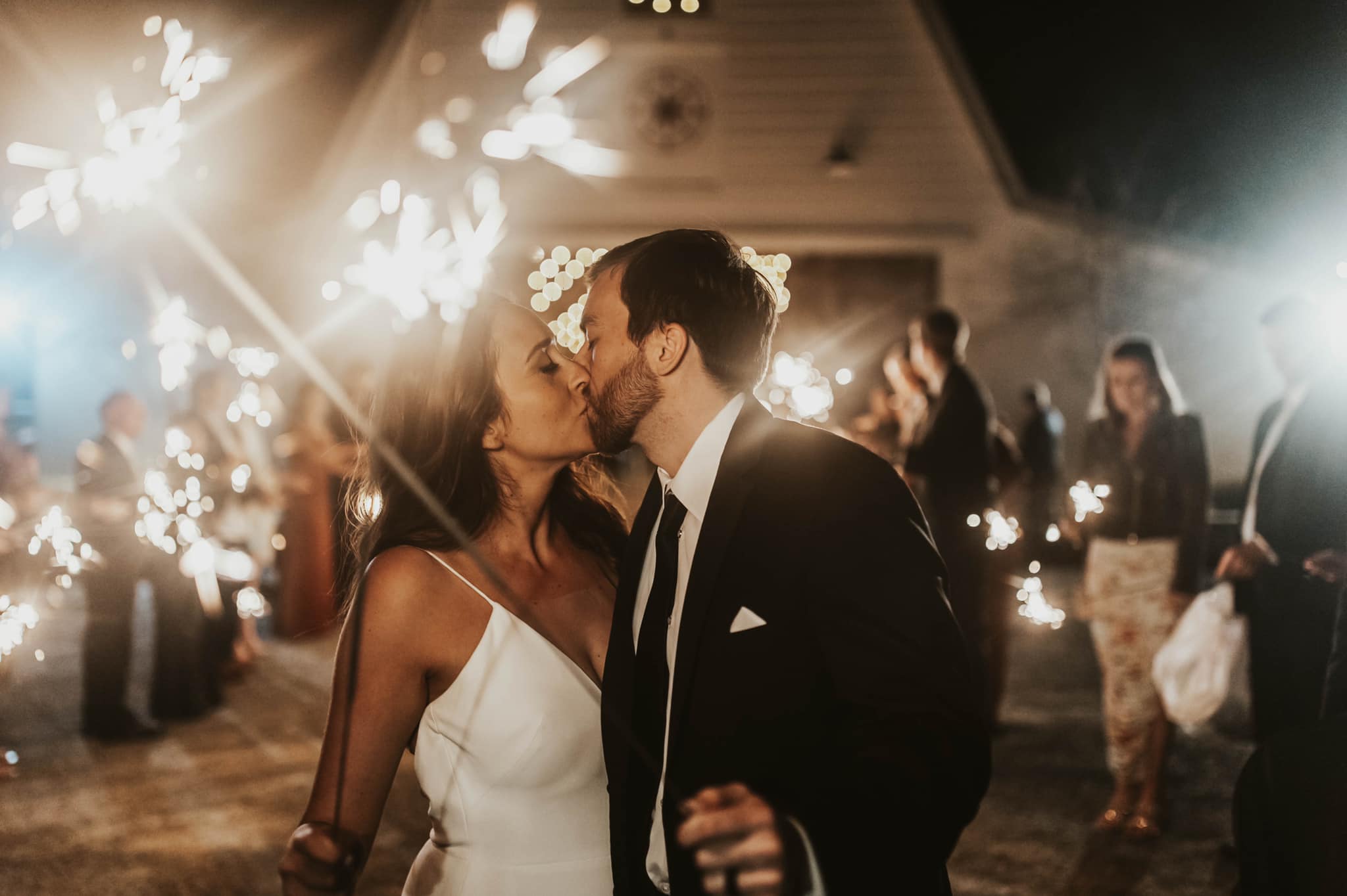 a newly wed couple kissing with the wedding party giving them a grand sparkler exit 