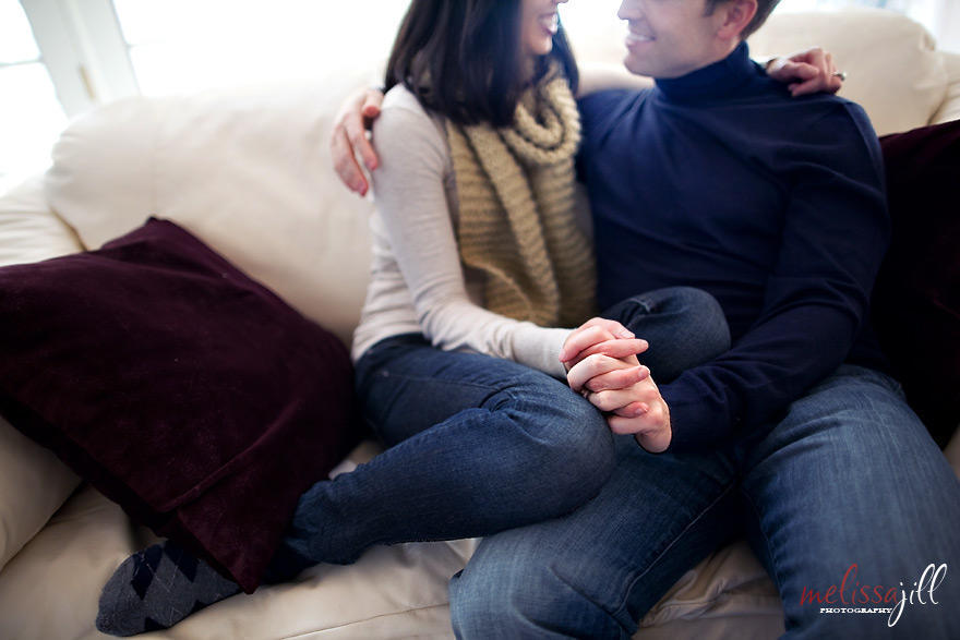 An engagement session photo indoors of the couple sitting on the couch and holding hands, with only the lower half of their faces in the photo.