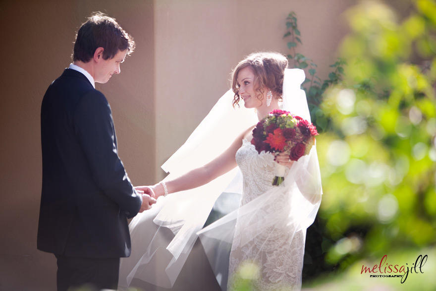 A wedding photography image with the bride and groom looking at one another, with the groom holding the bride's right hand, while her left hand holds her bouquet.