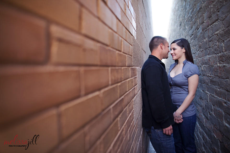 Couple in an engagement session in between two walls in an alley (ordinary places to take pictures near me/you) facing one another & holding one hand.