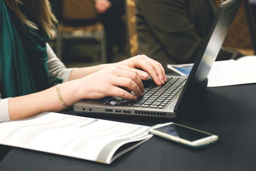 An image of a woman's hands, wearing a green scarf and off white sweater, as she types on a laptop sitting on a table with a pamphlet and phone next to the laptop.